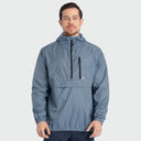 A man wearing a slate blue hooded anorak jacket with black zippers, standing against a light gray studio background.