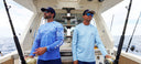 Two men on a boat wearing blue fishing shirts and hats, holding fishing rods.