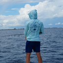 Person wearing a light blue hoodie with branding, standing on a boat over water with a cloudy sky.