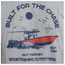 A close-up of a heather gray t-shirt featuring a vintage-style graphic print. The design shows a center console boat on waves under a sun and birds, with the text 'BUILT FOR THE CHASE' arched above and 'GUY HARVEY SPORTFISHING OUTFITTERS'