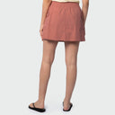 A model from the back wearing a dusty rose utility fishing skort with side pockets, paired with black flip-flops against a light gray studio background.