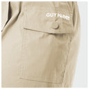 Close-up of a beige utility pocket on a pair of pants with 'GUY HARVEY' printed in white on the flap, featuring a matching button closure.