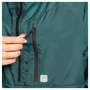 Close-up of a dark teal green lightweight jacket with a hand opening a black zippered pocket, showing the textured fabric and a small white brand tag.
