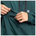 Close-up of a person wearing a dark teal green lightweight jacket, showing both hands opening a zippered pocket on the chest.
