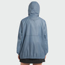 A person shown from the back wearing a hooded lightweight jacket in a muted dusty blue color, paired with a black skirt, against a light gray studio background.