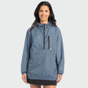 A woman wearing a slate blue hooded pack and stow jacket with black zipper details, standing against a light gray studio background.