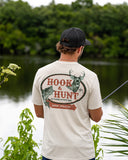 A man freshwater fishing while wearing a beige Guy Harvey HarborLite t-shirt with a Hook & Hunt themed graphic on the back.